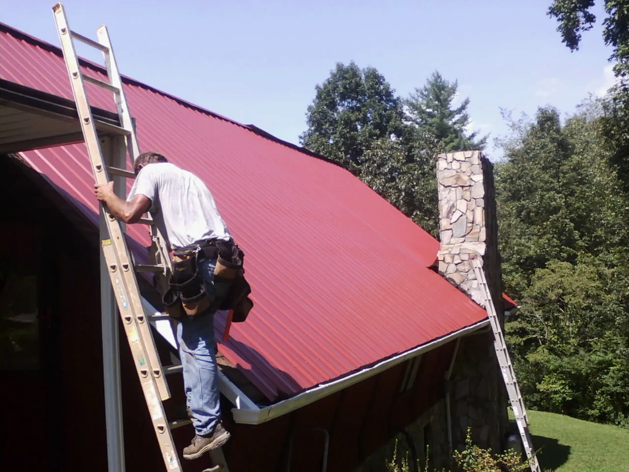 Roofer on a ladder beside a red metal roof and stone chimney — Soffit & Fascia Repair by Roofing Brothers in Gary